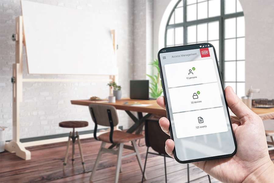 eine Hand, die ein Smartphone mit der ENiQ-App-Hauptbildschirmoberfläche vor einem kleinen Konferenzraum hält
