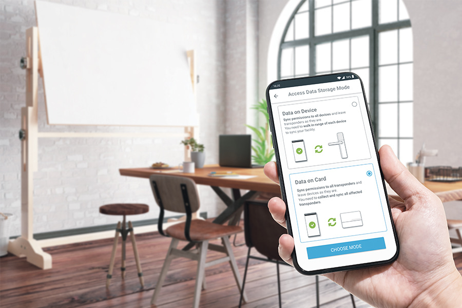 eine Hand, die ein Smartphone mit ENiQ-App-Programmiermethoden-Schnittstelle vor einem kleinen Konferenzraum hält