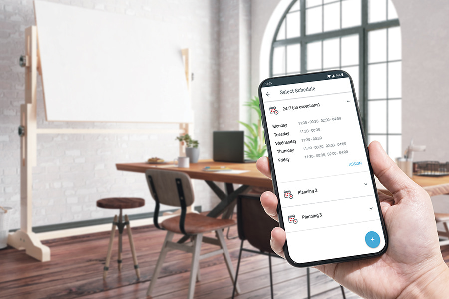 eine Hand hält ein Smartphone mit ENiQ App Wochenplanoberfläche vor einem kleinen Konferenzraum