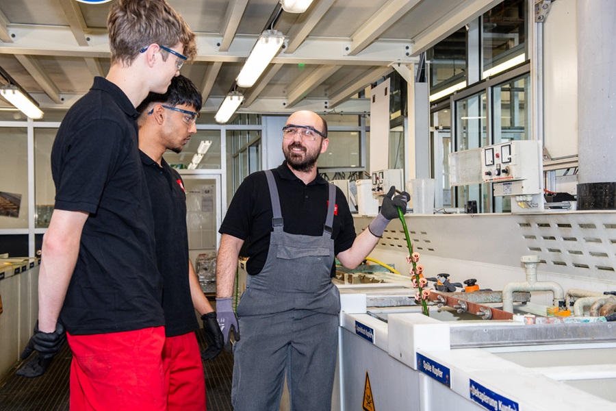 Apprentices for surface coating technology standing at an electroplating system while an instructor explains the coating process and lifts components from the bath.